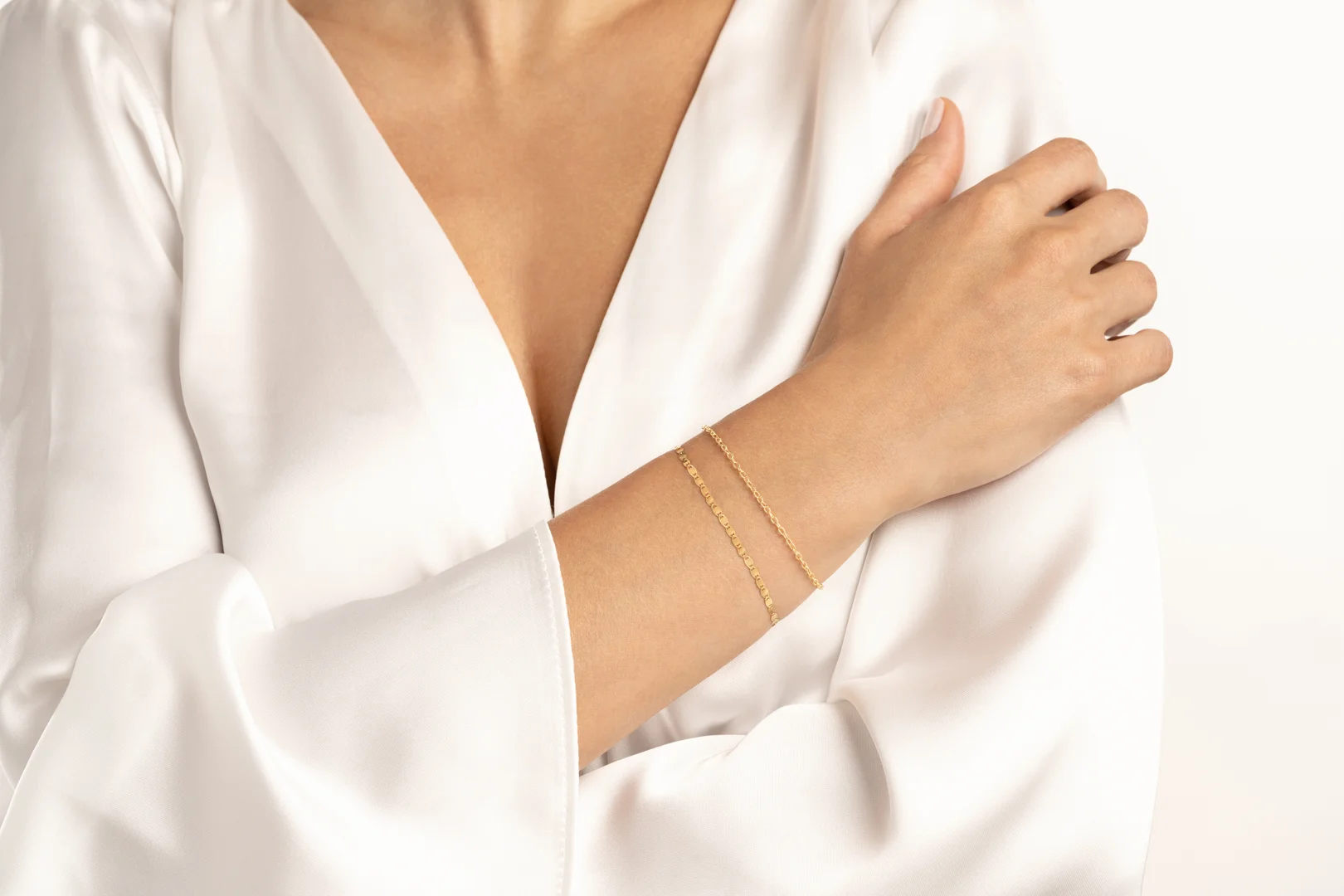 A close-up of a woman's arm draped across her body showing two 14 karat yellow gold permanent bracelets on her wrist and a loosely draping white blouse or dress.