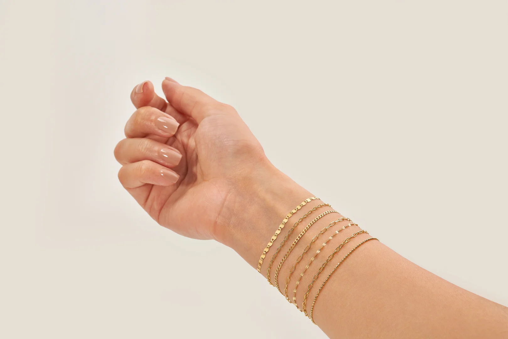 A close-up of a woman's wrist that shows seven different 14 karat yellow gold permanent bracelets on an isolated beige background.