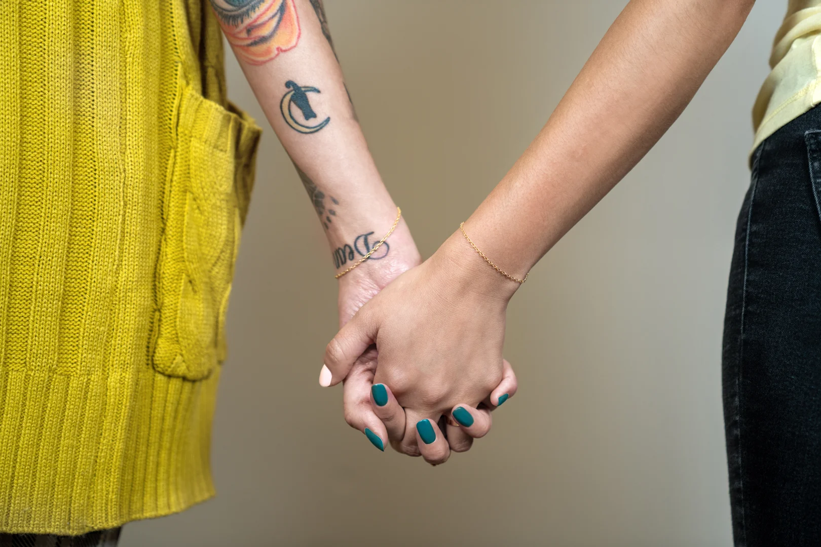 A close-up of two women holding hands with their yellow gold permanent jewelry bracelets visible on their wrists.