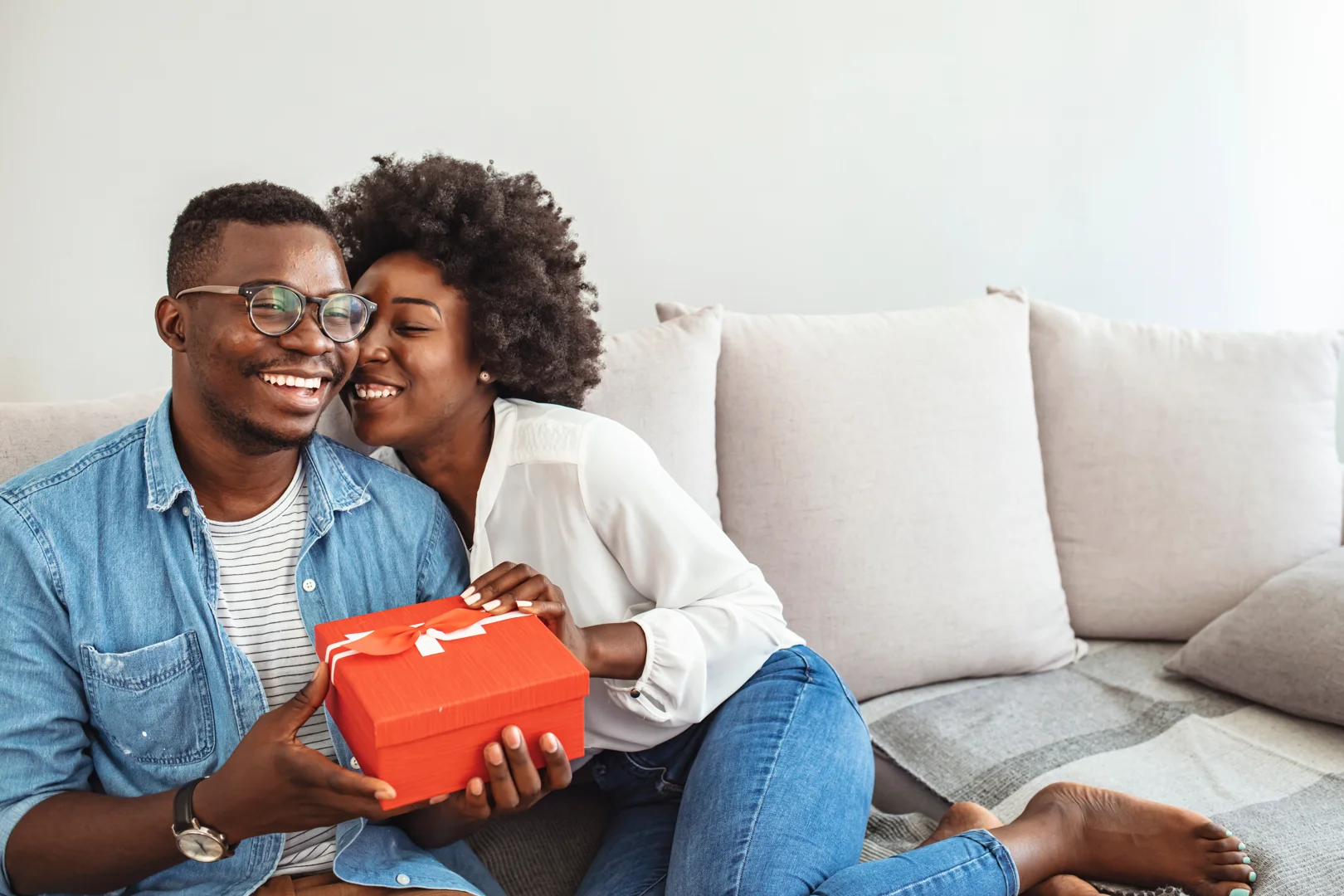 A young happy and smiling couple sit on a white beige couch as the woman gently pushes a 12th anniversary gift box towards the man.