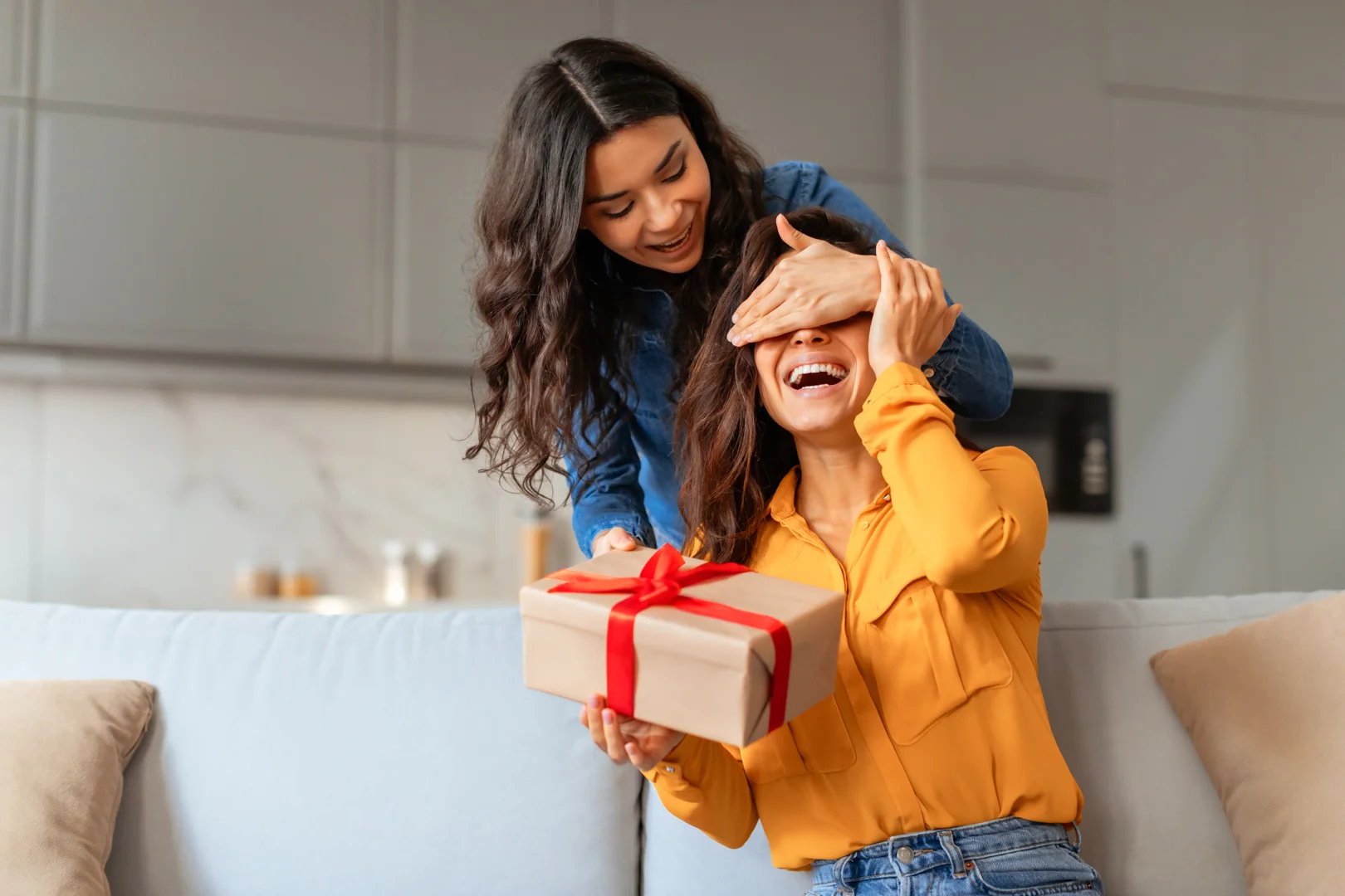 A woman sits on a couch with a 12th anniversary gift box in one hand and her partner's hand over her eyes to surprise her with her gift.