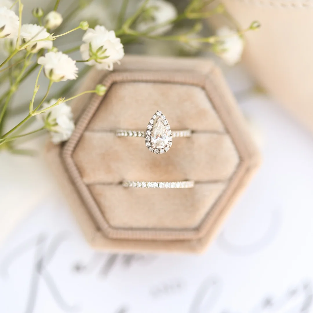 A diamond halo engagement ring and classic wedding band bridal ring set in a pale pink ring box with white flowers and blurred writing in the background.