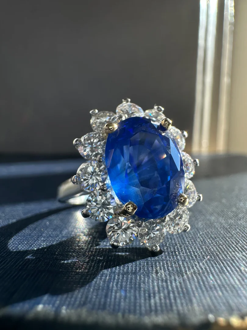 A custom-designed white gold oval blue sapphire and diamond halo ring resting on a textured navy blue surface with sunlight coming in from the right side.