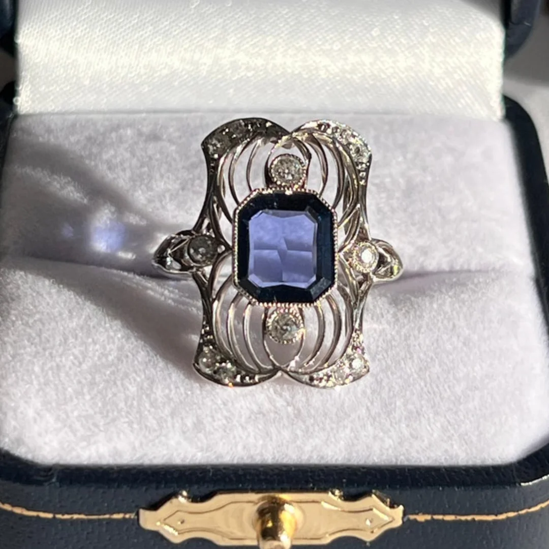 user-inserted A vintage blue sapphire and diamond filigree ring resting on the white velvet interior of a jewelry box with dark blue and gold accents on the outside.