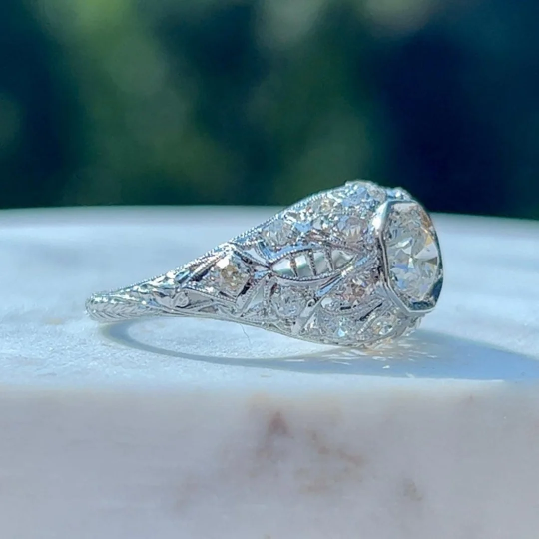 user-inserted An antique Art Deco engagement ring with an old mine-cut center diamond and delicate filigree and metalwork. The ring rests on a white marble surface with blurred trees behind it.