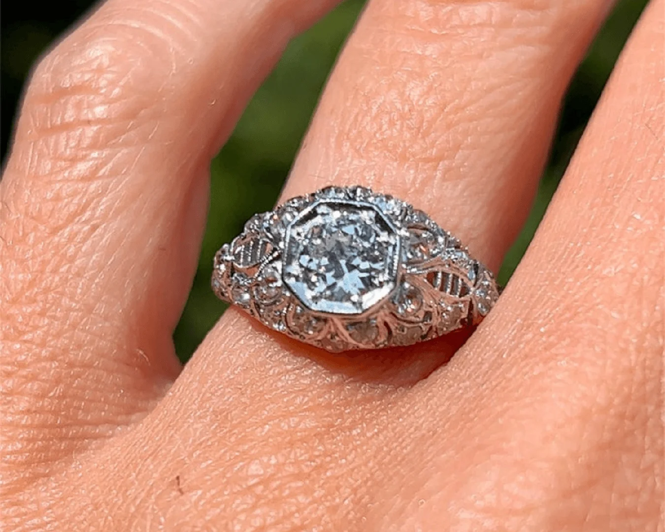 An antique Art Deco old European-cut diamond engagement ring on a woman's left ring finger with blurred green grass in the background.