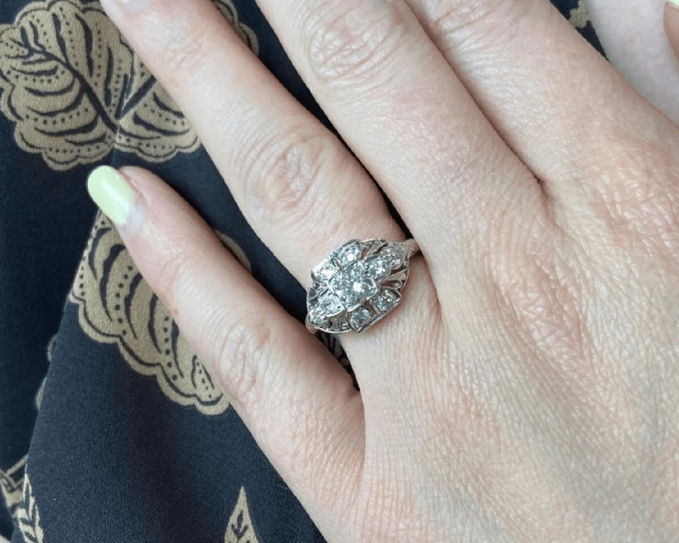 An antique Art Deco diamond engagement ring on a woman's hand who has pale yellow nail polish with her hand placed next to dark navy blue fabric with gold leaf accents.