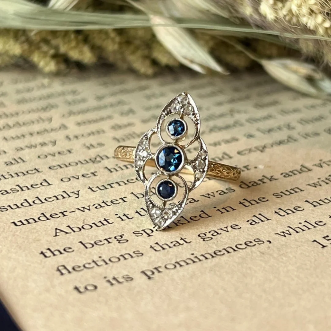 user-inserted An antique Edwardian navette ring crafted from yellow gold and platinum with blue sapphires and diamonds. The ring rests on the open page of a book with blurred plants in the background.