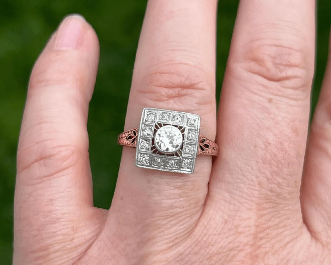 An antique Edwardian diamond ring on a woman's left ring finger with blurred green grass in the background.