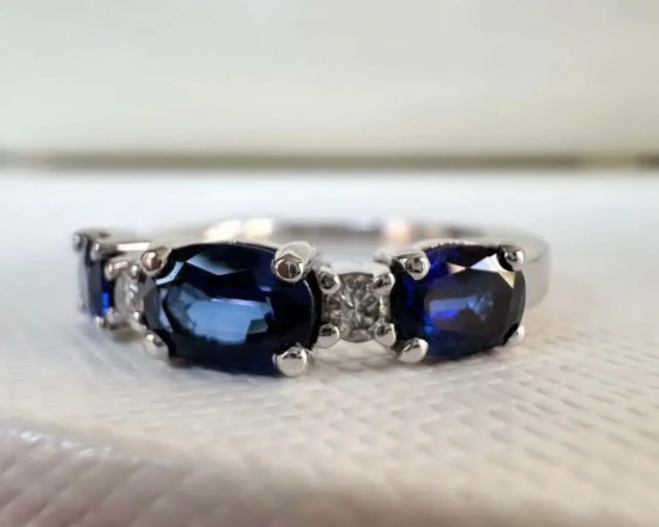 A final product of a custom jewelry design project for a white gold blue sapphire and diamond anniversary band resting on soft, textured, white fabric.