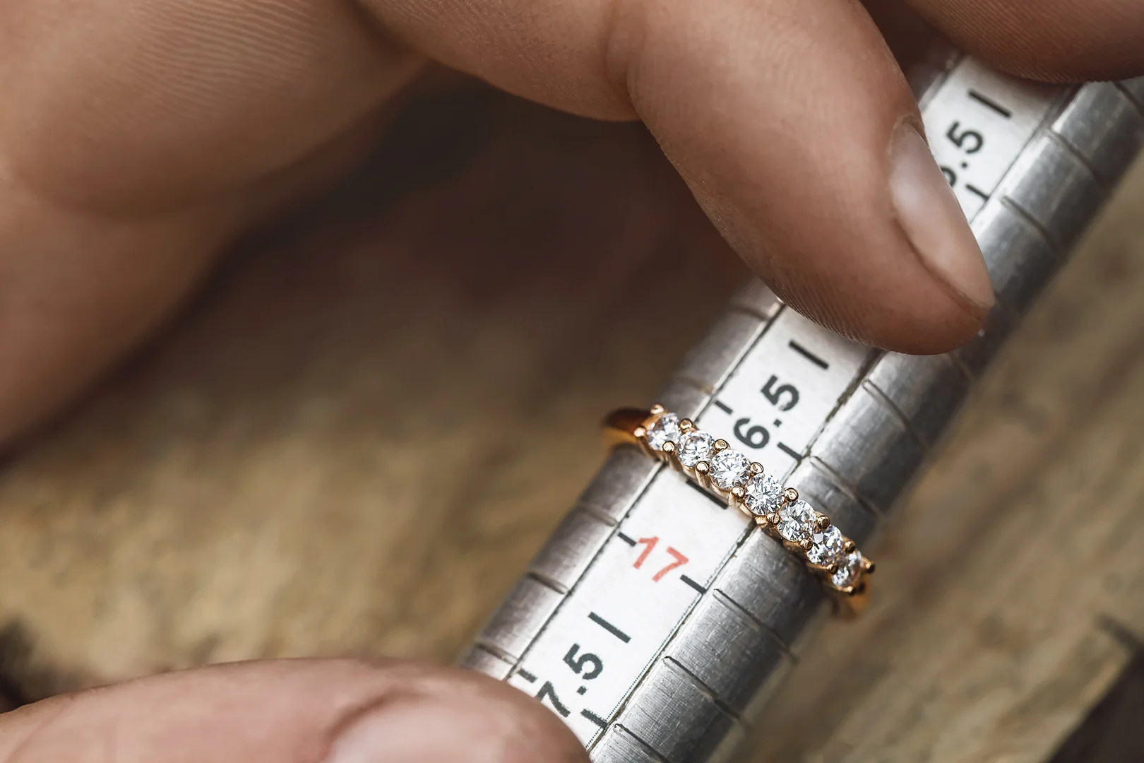 A diamond band on a jeweler's mandrel in a person's hand with a blurred wood grain surface in the background.