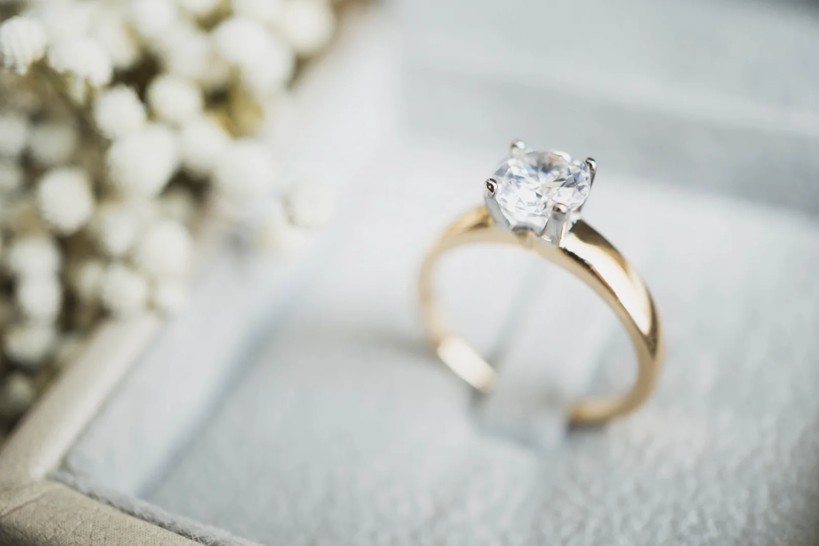 user-inserted A 14-karat yellow gold and white gold four-prong round brilliant-cut diamond solitaire engagement ring secured in a luxury jewelry box with flowers blurred to the side.