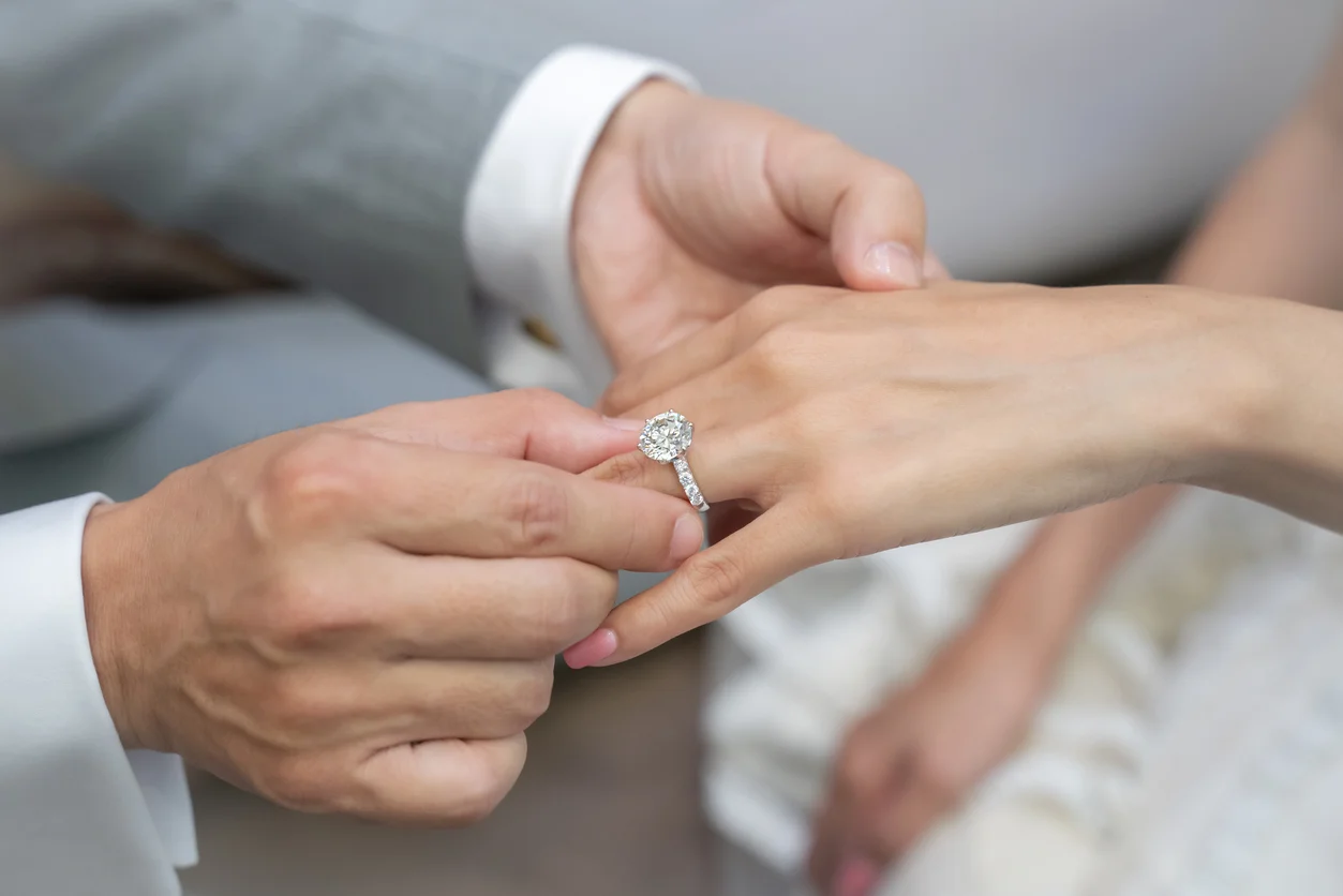 user-inserted A close-up image of a man's hand gently placing a diamond engagement ring on a woman's ring finger.