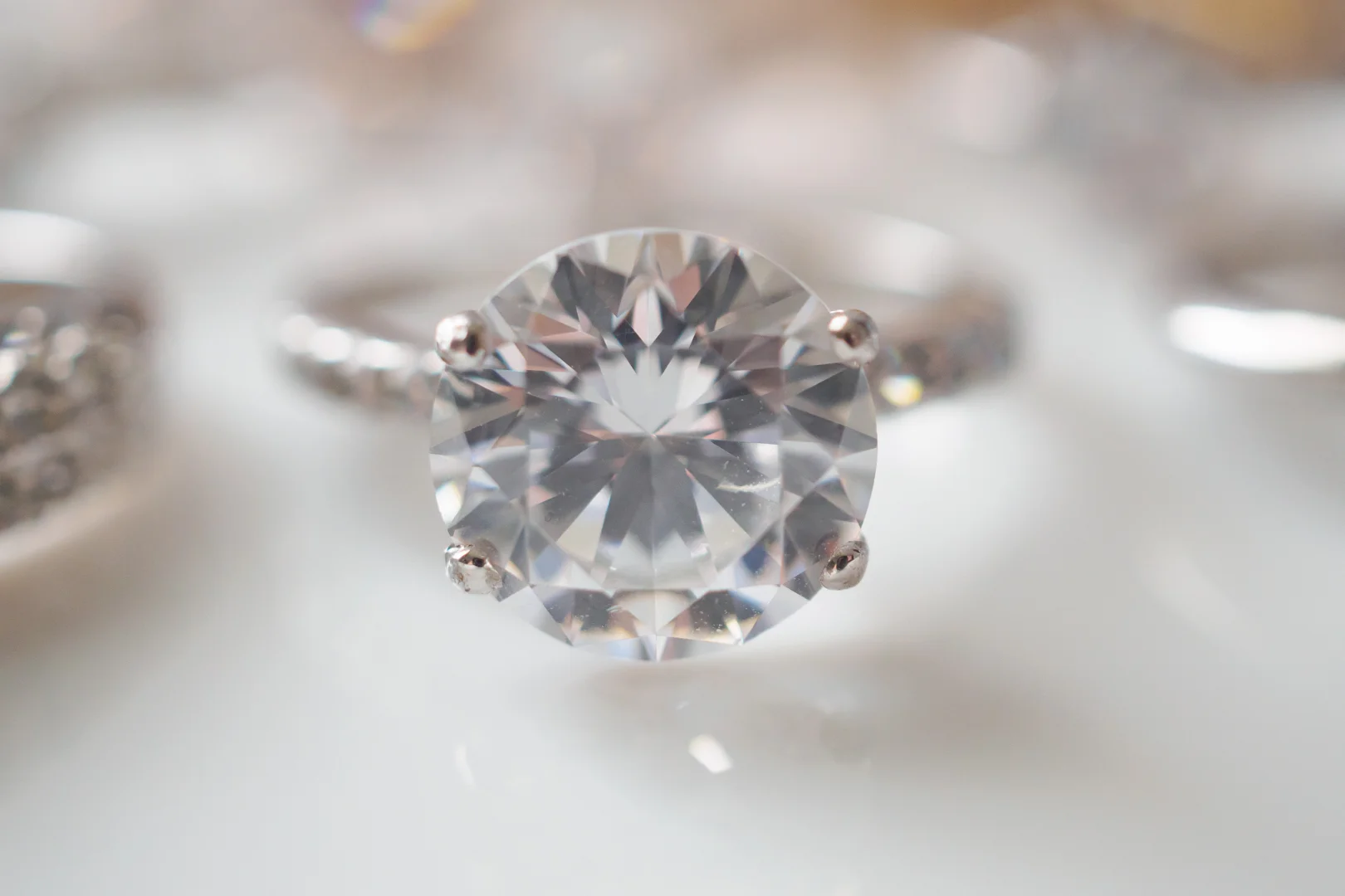 user-inserted A 14-karat white gold four-prong round brilliant-cut diamond solitaire engagement ring on a white surface with other blurred engagement rings nearby.