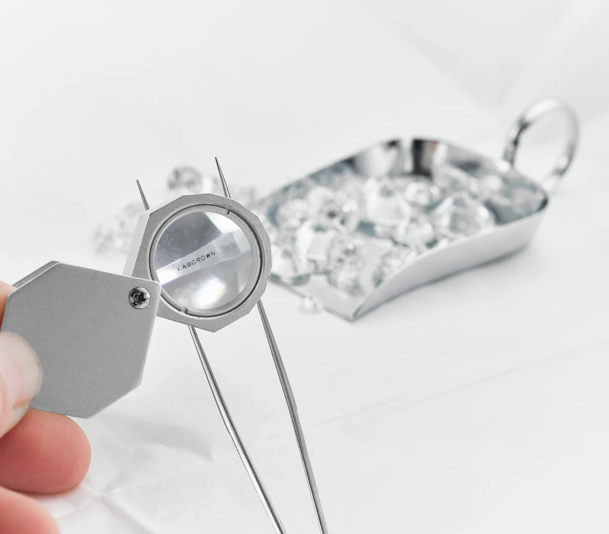 A jeweler's loupe focuses on a slip of paper that says "LAB GROWN" being held by jeweler's tweezers with a small container of lab-grown diamonds in the background
