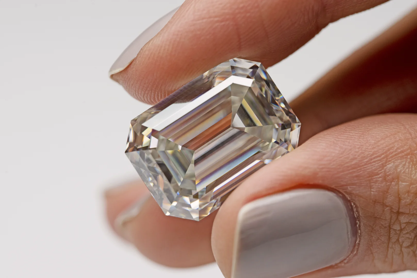 user-inserted A close-up of a loose emerald-cut diamond being held between a woman's thumb and ring finger on a greyish beige background.