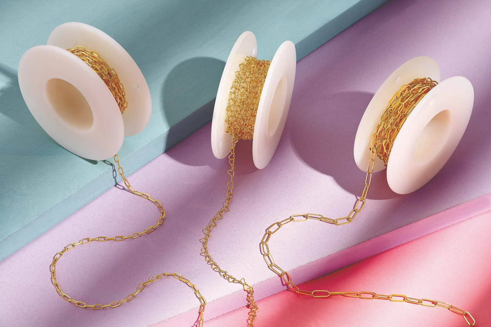 Three spools of permanent jewelry bracelet chains on colorful surfaces of turquoise, purple, and pink.
