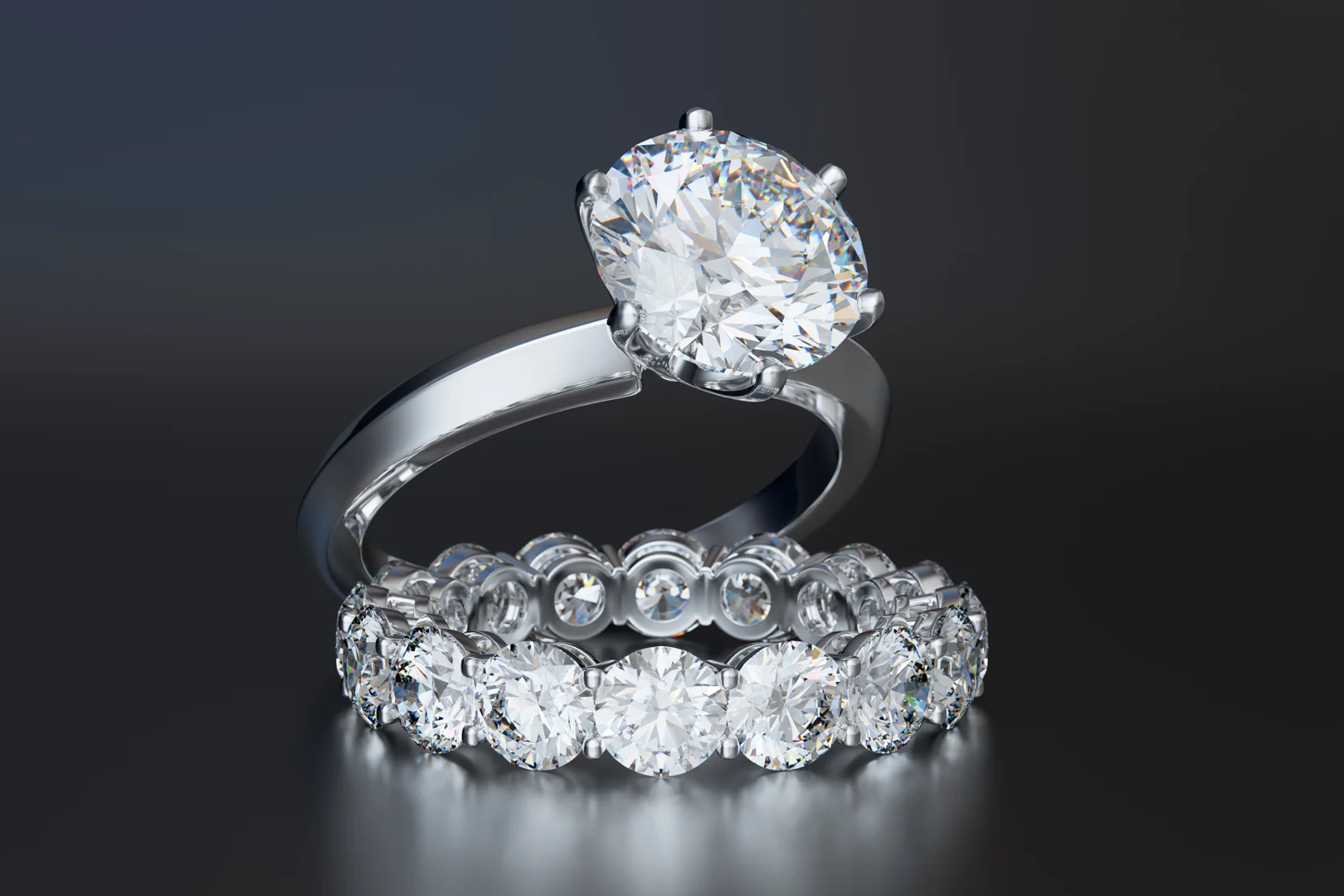 A diamond bridal ring set containing a six-prong diamond solitaire engagement ring with a knife-edge band and a round diamond eternity wedding band on a reflective dark grey background.