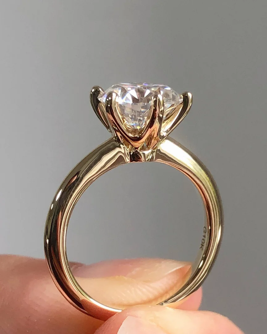 A six-prong diamond solitaire engagement ring with an open basket design being held by a person's thumb and pointer fingers.