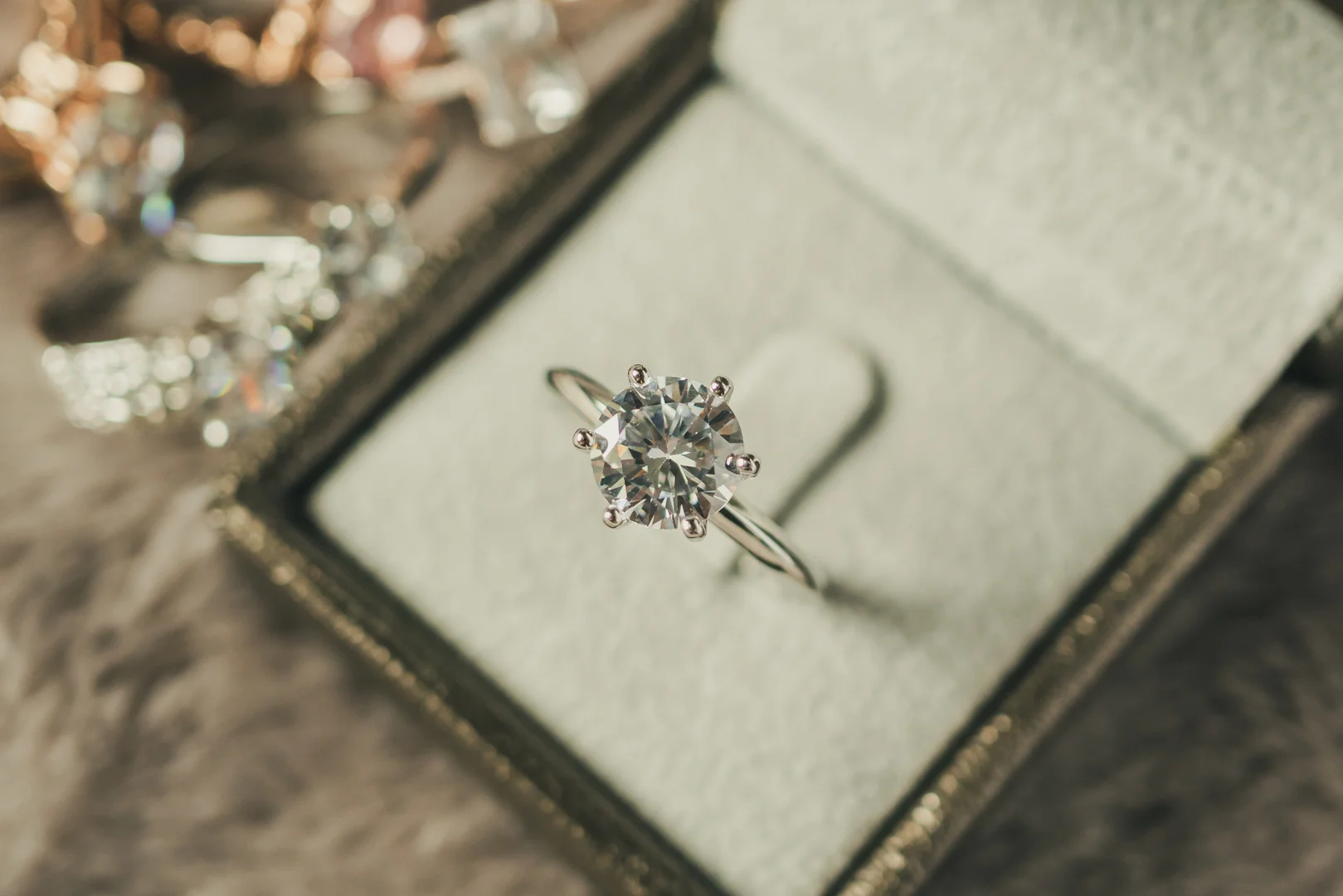 A white gold six-prong diamond solitaire engagement ring in a beige jewelry box with a blurred background.