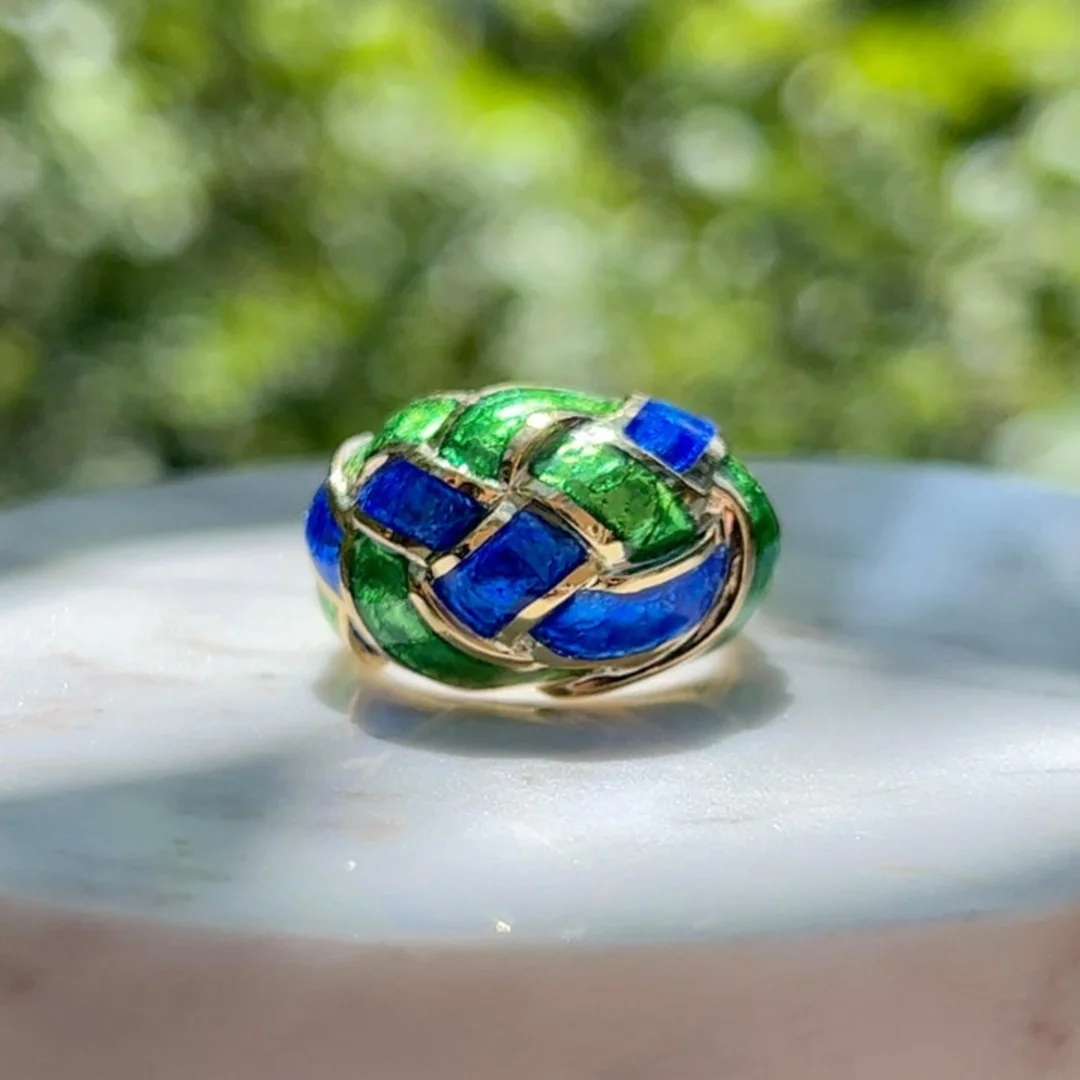 user-inserted A vintage Tiffany & Co. ring designed by Jean Schlumberger, crafted from yellow gold, containing a blue and green enamel weave pattern. The ring rests on white marble, with blurred trees behind.