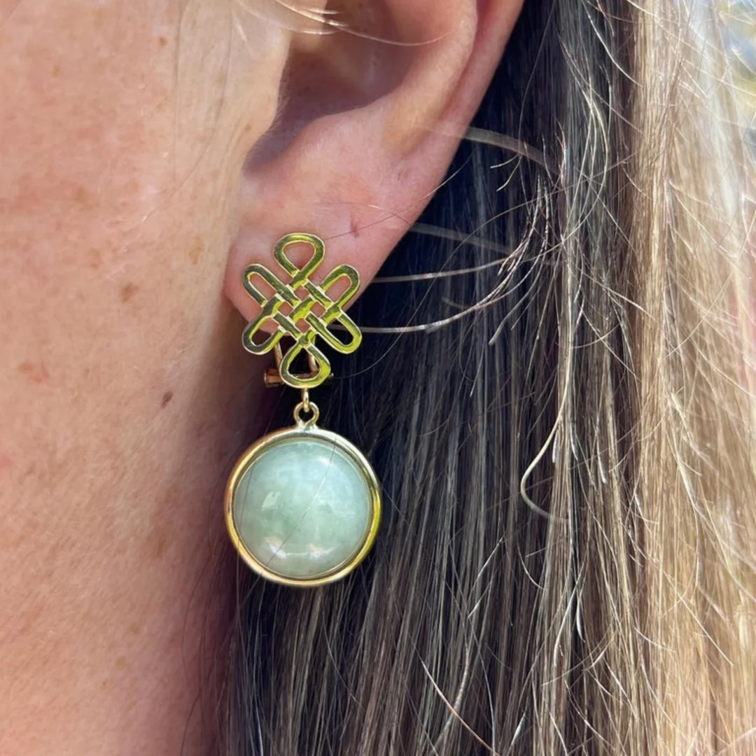 A close-up image of a vintage yellow gold jade drop earring on a woman's ear with long brown hair behind it.