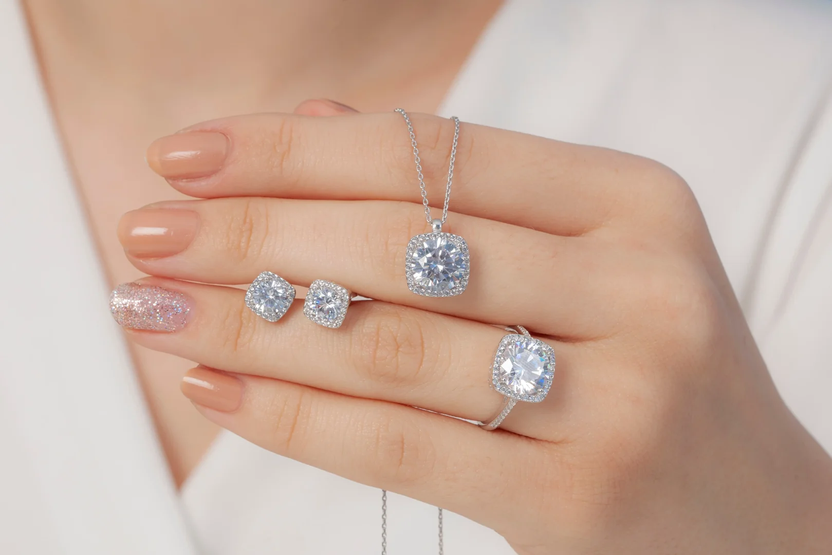 A close-up of a woman's hand holding up a collection of diamond jewelry, including diamond halo stud earrings, a diamond halo pendant necklace, and a diamond halo ring.