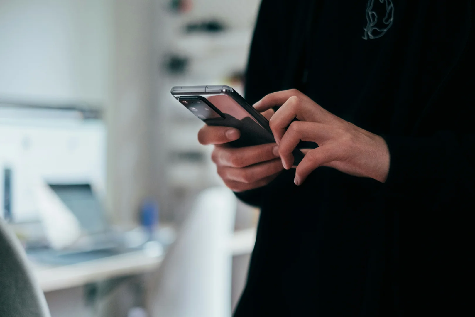 A person wearing a black shirt uses a smartphone with a large rear camera array, blurred office workspace in background.