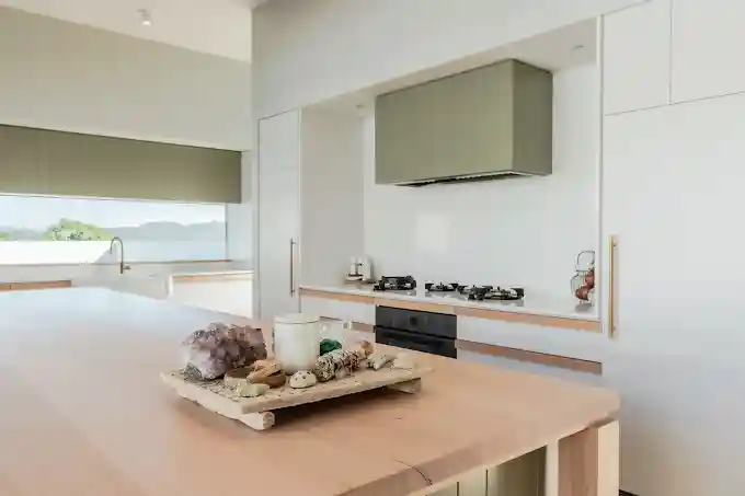 Minimalist modern kitchen with a large wooden island in the foreground, white cabinetry, built-in stovetop with a grey range hood, and a window with a view of greenery and hills. Minimalist modern kitchen with a large wooden island in the foreground, white cabinetry, built-in stovetop with a grey range hood, and a window with a view of greenery and hills.