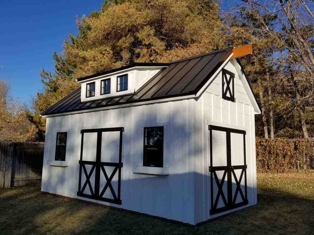 A barn-style shed with a white exterior, black trim, and a metal roof, surrounded by trees.