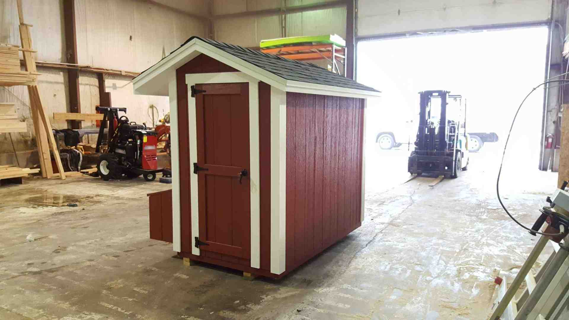 A small wooden chicken coop with red and white siding, partially built in a workshop with a forklift nearby.