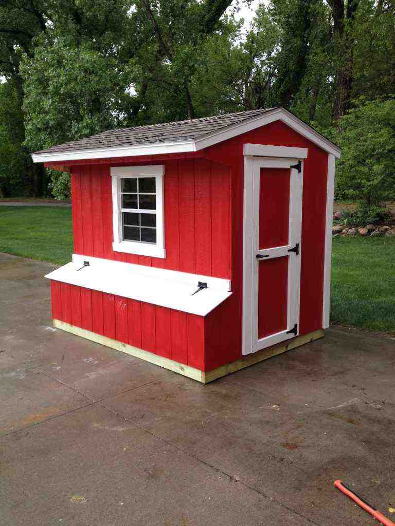 A small, red chicken coop with white trim, featuring a window and door, placed on a concrete surface.