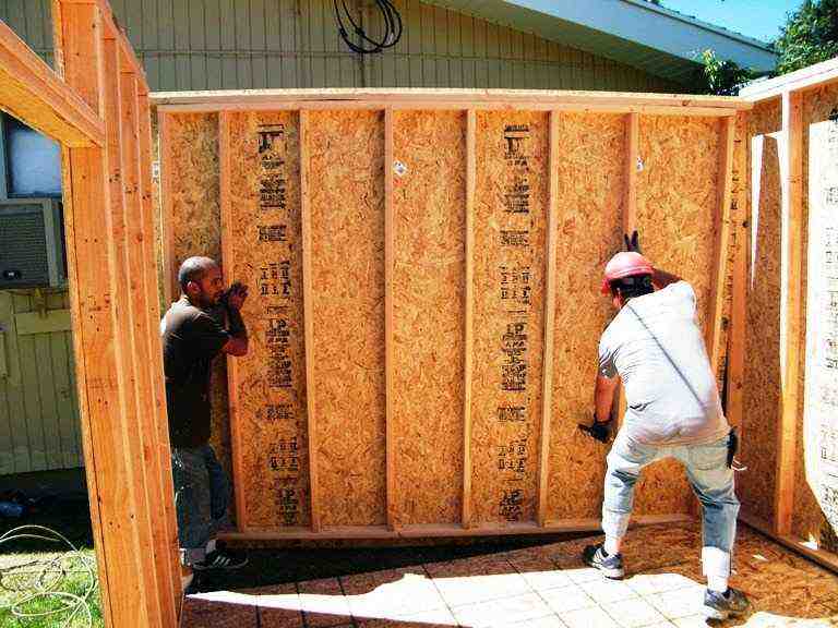 Two workers lifting and positioning the wall frame of a DIY storage shed, an important step in building your own shed.