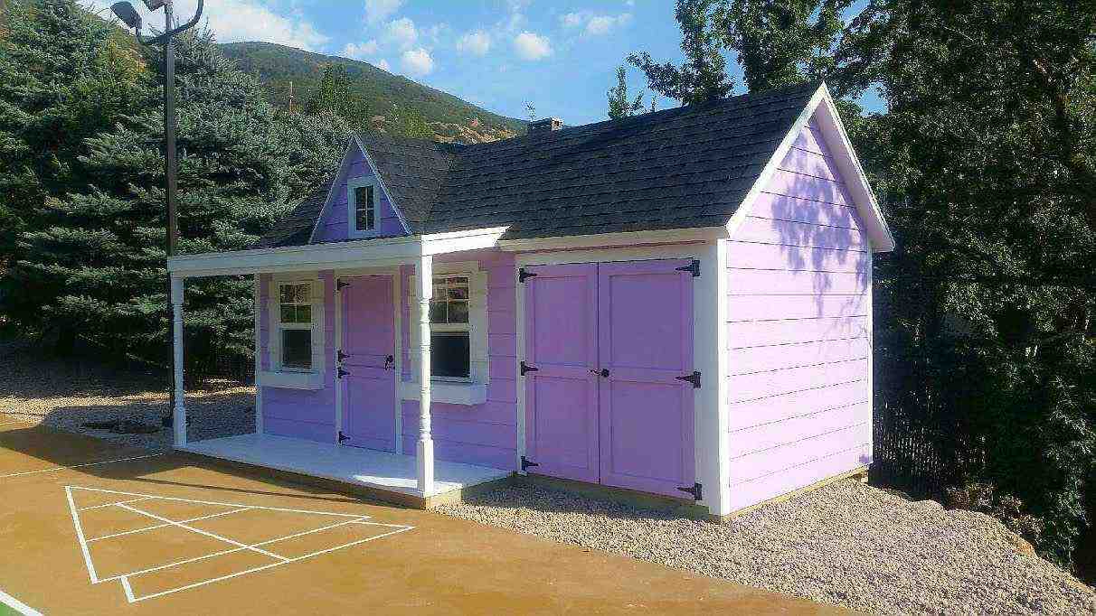 Lavender playhouse-style storage shed for sale in Utah with porch, double doors, and charming design.