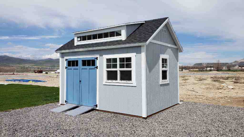 Light blue storage shed with double doors and windows on a gravel pad. Focus: storage shed.