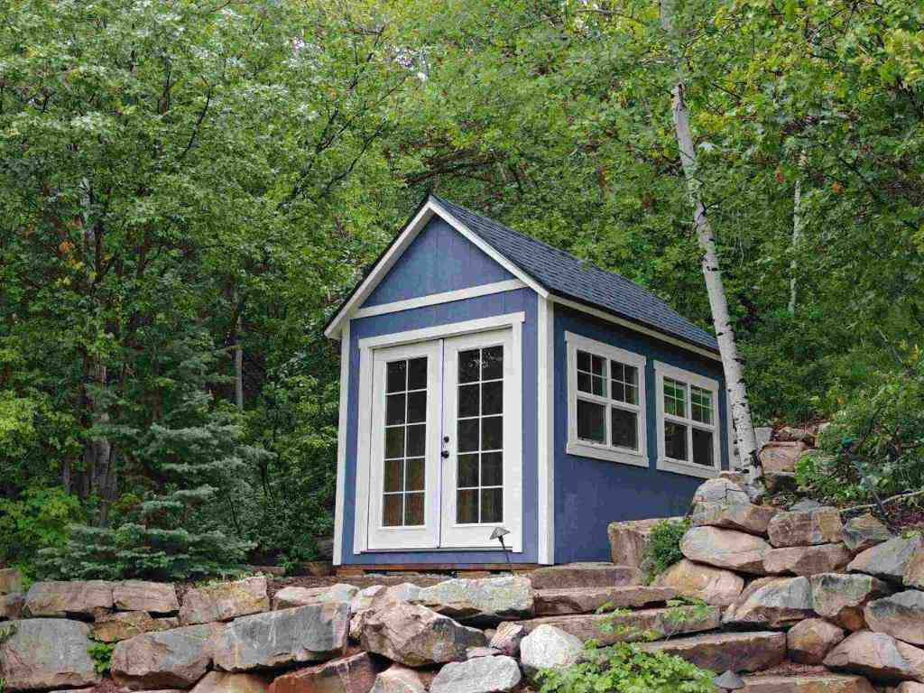 Blue storage shed with patio doors and large windows on a sloped landscape. Focus: storage shed.