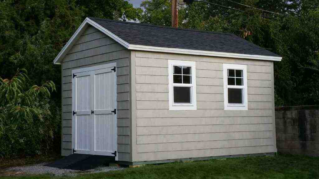 A backyard shed featuring a gray exterior with double doors and windows, surrounded by greenery.