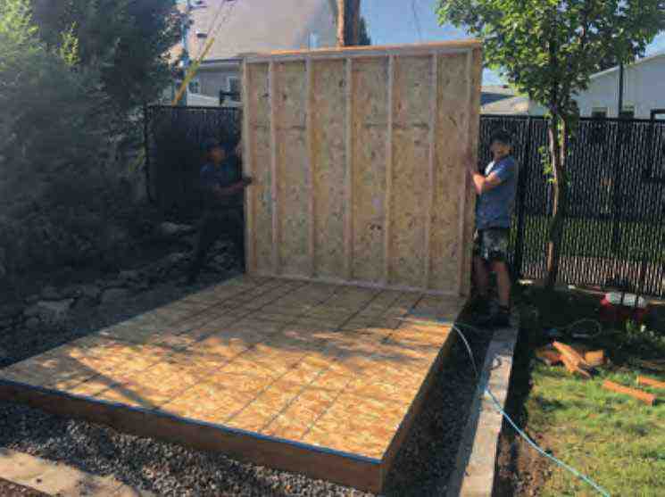 Installing Shed Walls During Assembly Two workers lifting framed wall panel during shed kit assembly on leveled wood floor platform in backyard construction site.