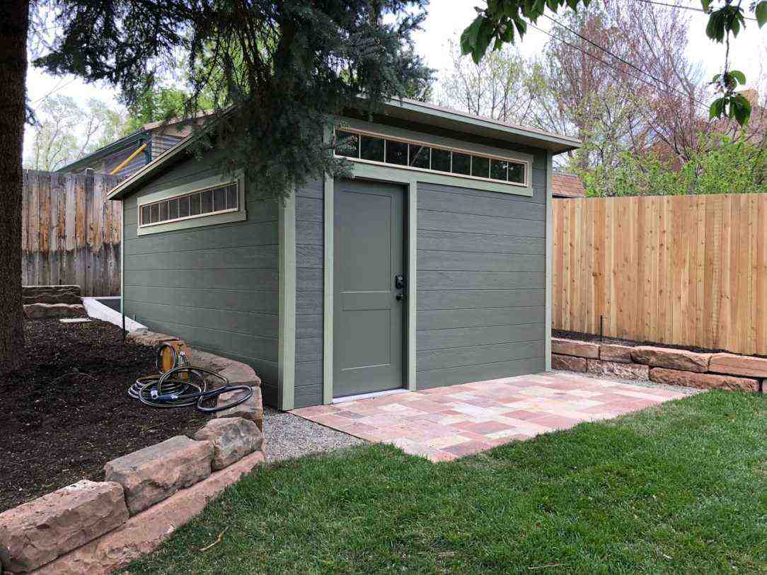 A lean-to shed with a green exterior, a door, windows, and a stone pathway, surrounded by greenery.