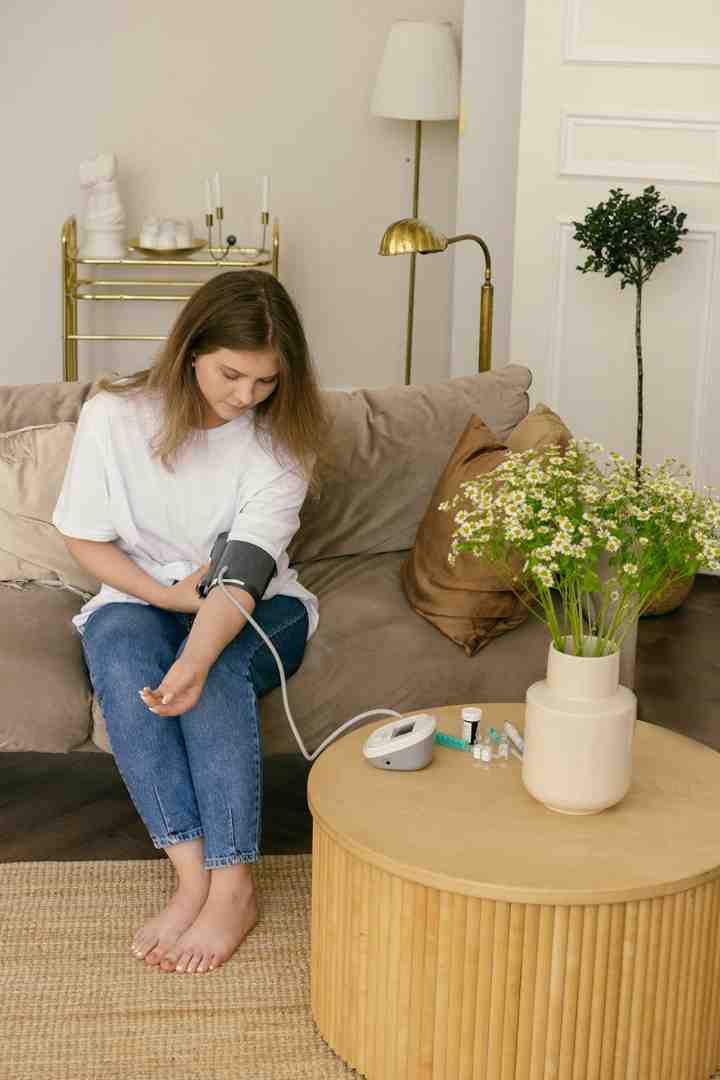 Woman in White Long Sleeve Shirt and Blue Denim Jeans Sitting on Brown Sofa with blood pressure monitor