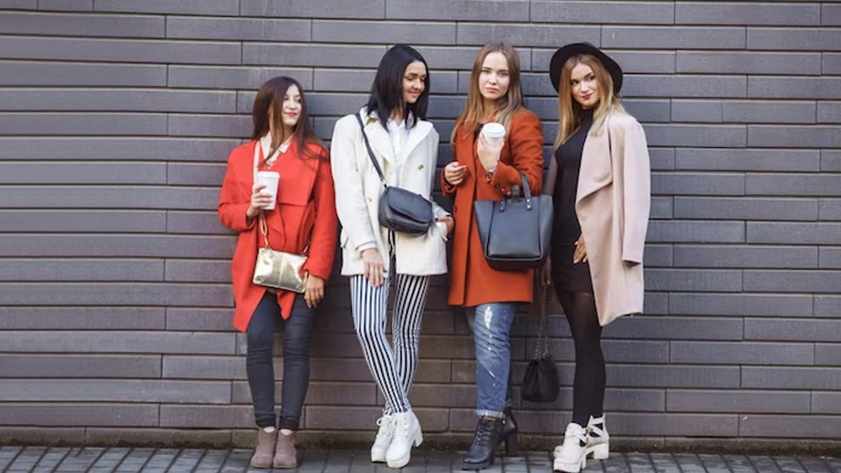 Four stylish women standing by a brick wall in trendy outfits, holding coffee cups and handbags, showcasing modern fashion