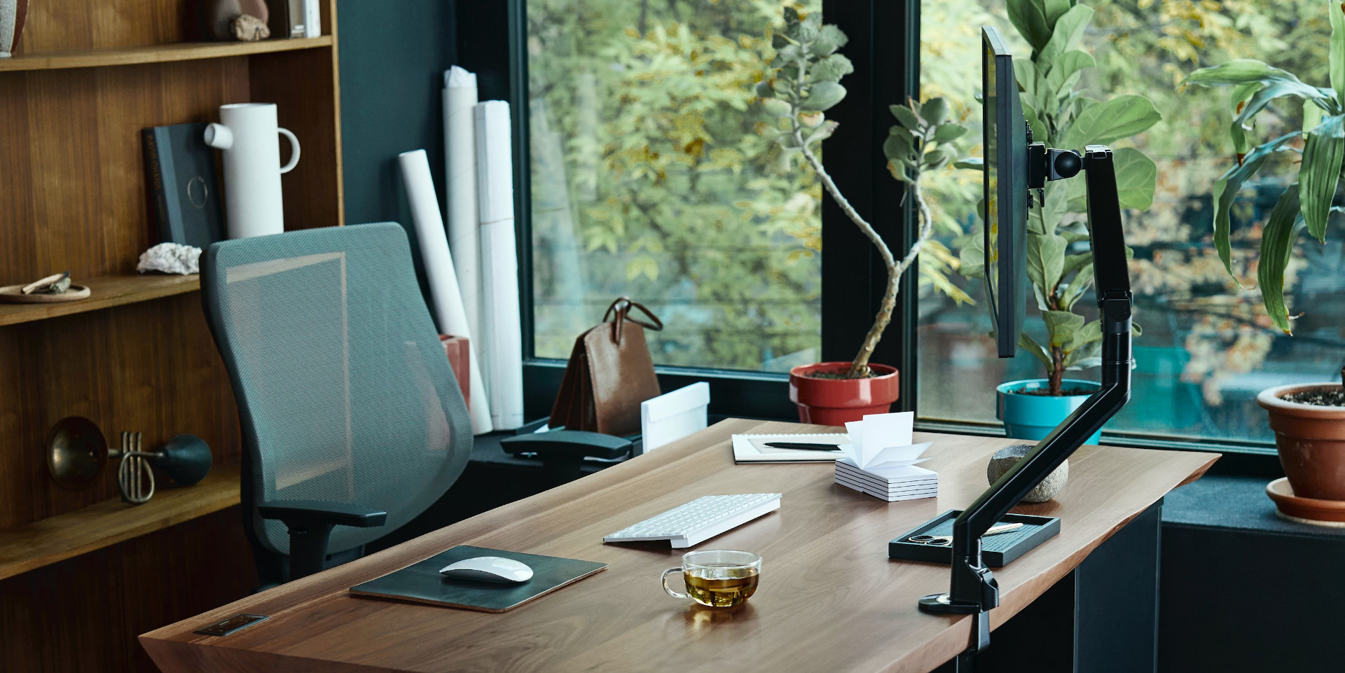 Home office featuring a desk beside a window overlooking some trees, with potted plants sitting on the windowsill. Facing the desk is an office chair, behind which is a sparsely decorated bookshelf. on the desk is a keyboard and mouse, computer screen, pen and paper, and a cup of tea.