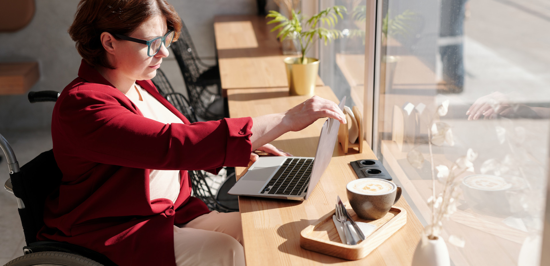 Woman in a red jacket sitting at a coffee shop bar in a wheelchair, facing out the window and adjusting the screen on her laptop.