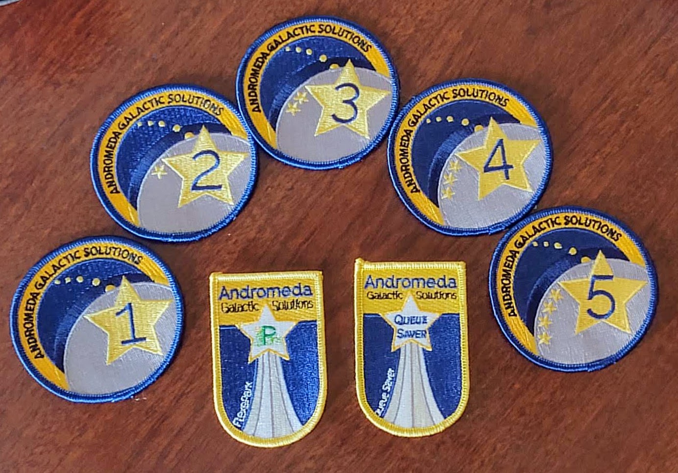 Mission Patches for Andromeda Galactic Solutions. Annual patches are round and show from 1 to 5 stars to represent the number of years with the company. There's one large star with the number in it, then smaller star trailing behind it. There are also two patches shaped like a shield. Each has a shooting star with a product logo in the middle, and the stars tail makes it look like it's shooting up from beneath as a form of "launch" effect.