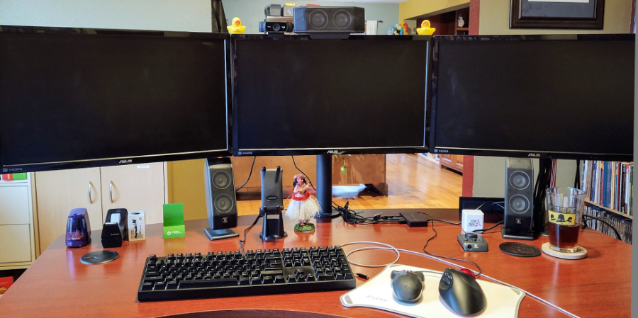 Photo of Michael's desk showing three HD screens side-by-side on a single mount, with keyboard and mouse in front and speakers on the sides. Other office decorations line the desk.