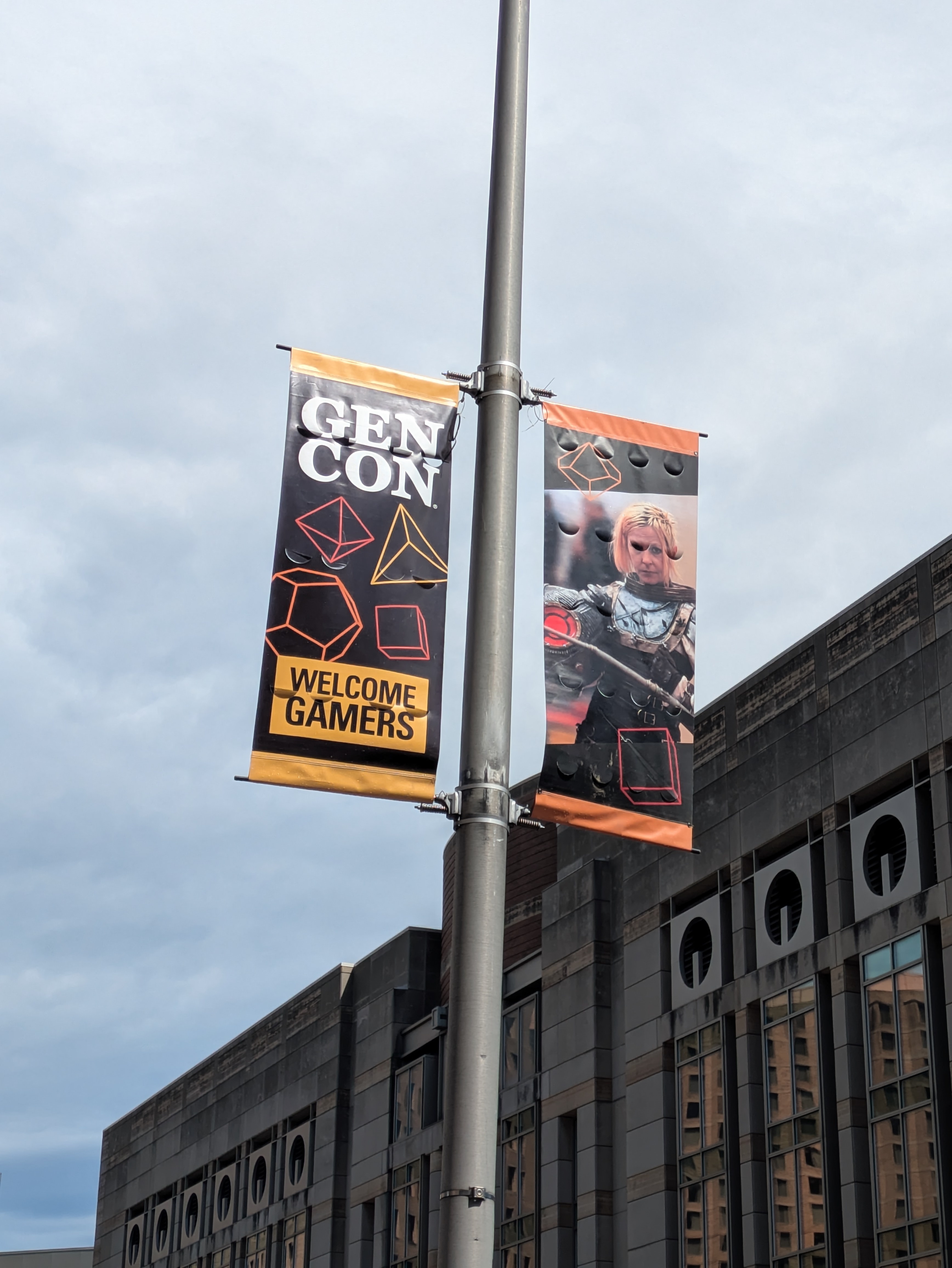 Banners hanging from a light pole in downtown Indianapolis. One has a picture of a cosplayer in a suit of armor, and the other says Gen Con above  outlines of polyhedral dice, and Welcome Gamers.