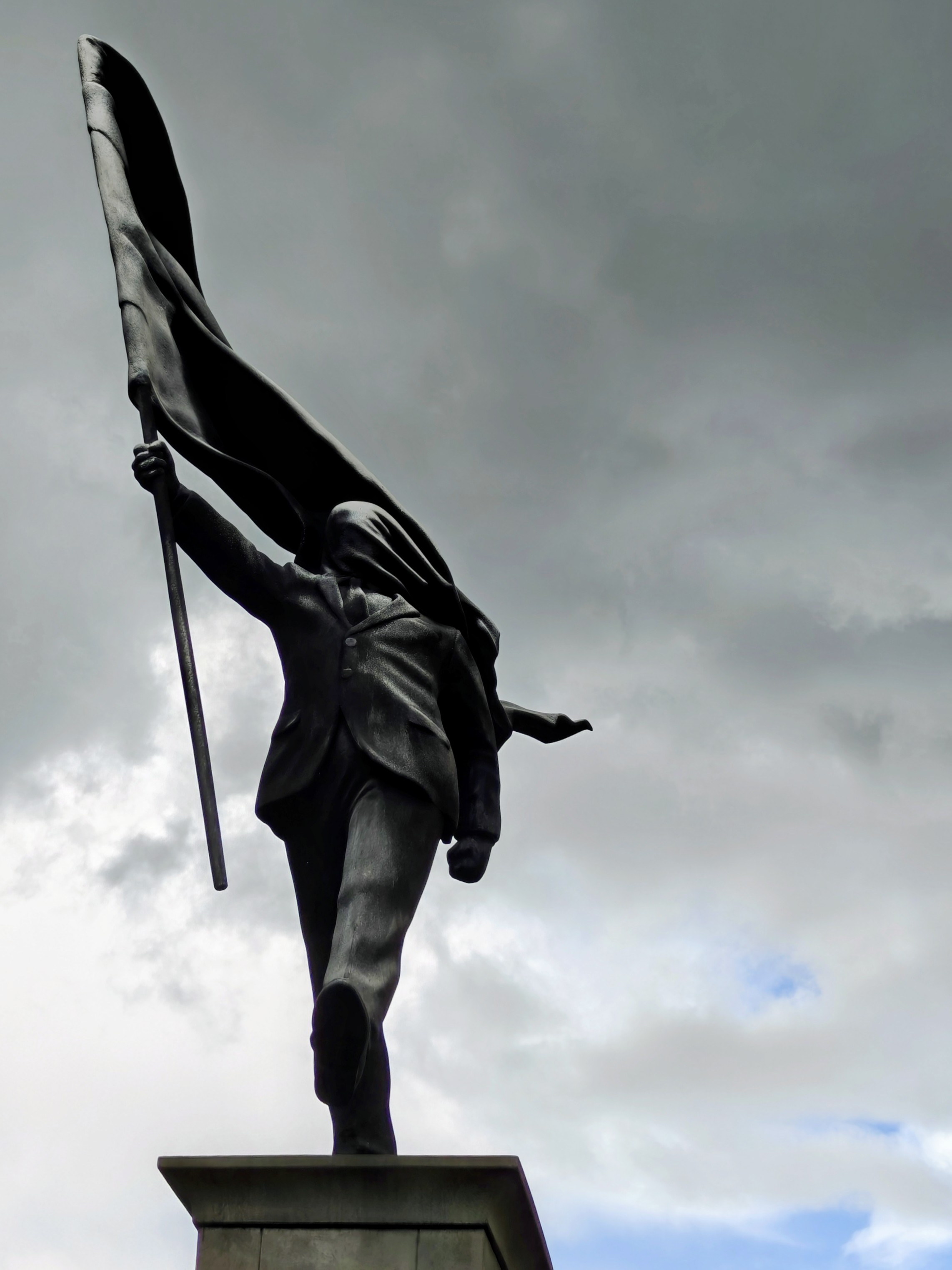 Photo of the latest Banksy sculpture at Waterloo Place in London. The perspective is looking up at the statue cast in shadow against a stormy sky. The statue depicts a suited man walking forward off a plinth while carrying a flag that covers his face.