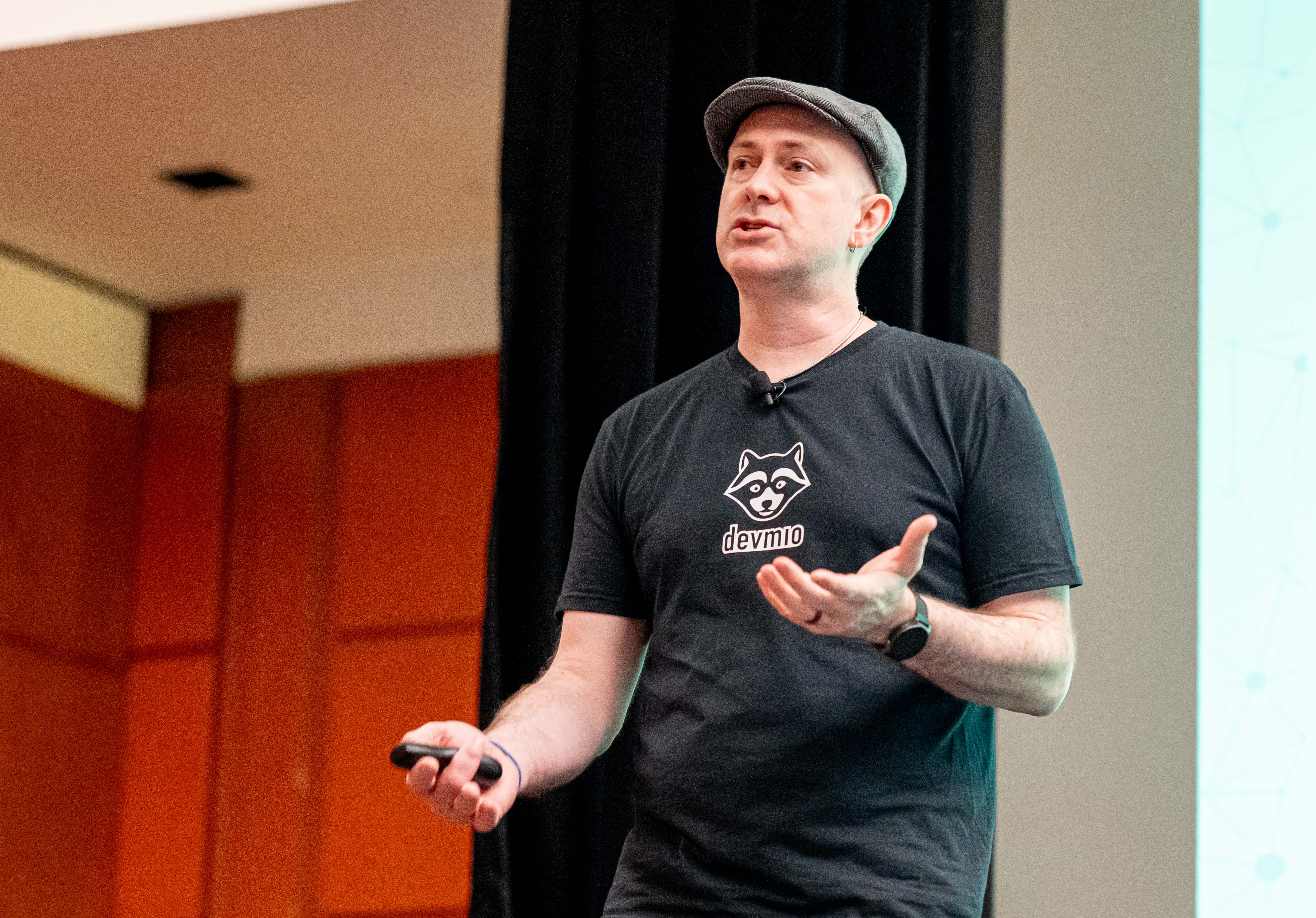 A close shot of Michael Dowden, in a black devmio shirt, gesturing and speaking in front of an audience.