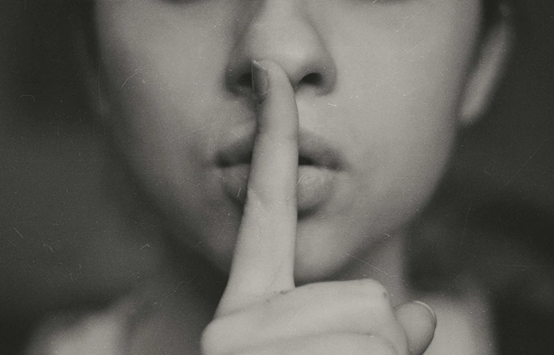 A woman's face with a finger in front of her mouth indicating that one should be quiet or not make noise