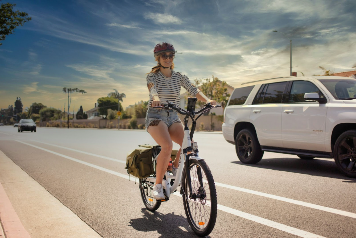 Woman wearing shorts, long sleeves, and a helmet while riding a bike along a highway in the bike lane. She is being passed on the left by a large, white SUV. The sky is sunny and slightly overcast with visible clouds. The far side of the road is bordered by a wall with houses behind it. The  woman is smiling while she rides and her posture is mostly vertical with negligible forward lean. The bike itself has two canvas bags on the back for storage.